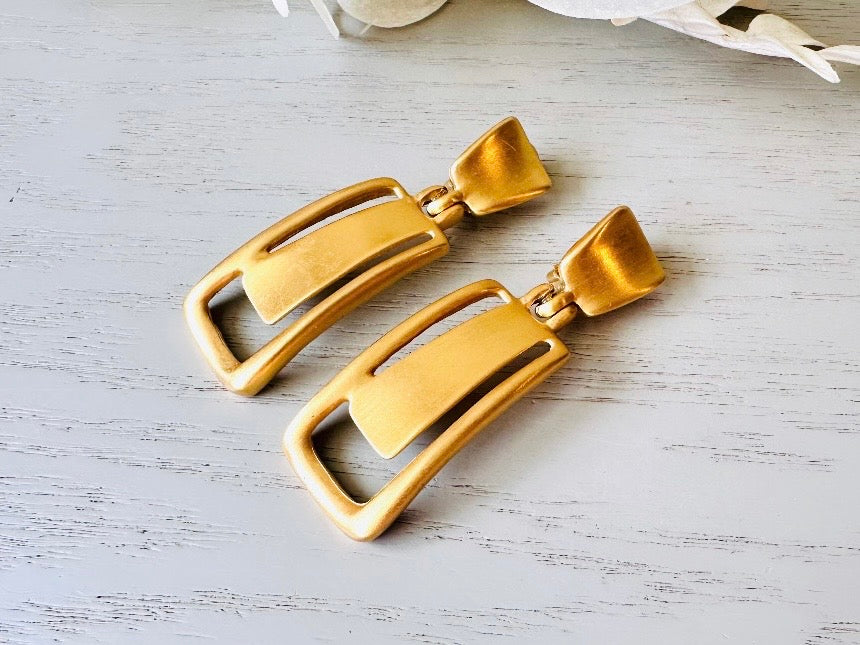 These Anne Klein vintage matte gold designer earrings are spectacular. Full of 1980s bold glamour, these clip ons have a satin gold finish and a geometric design. These vintage dangle earrings are bold and elegant, with curved square posts and rectangular drops with a cut out hollow border. These are big and incredibly well made in a satin matte gold finish. Clip on style, easy to wear and in great vintage condition. Highly sought after designer vintage.