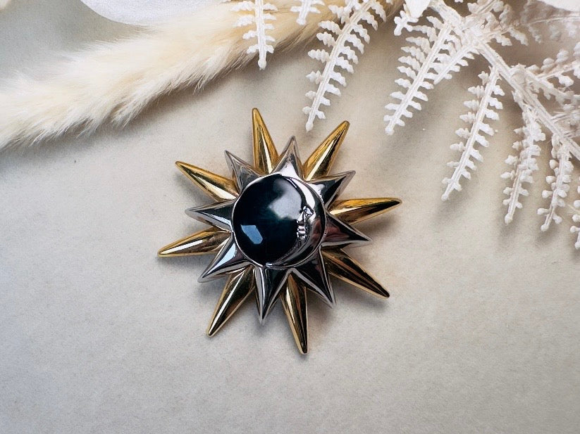 A starburst brooch featuring a central glossy black oval stone surrounded by alternating silver and gold spikes, displayed on a neutral beige surface with decorative white faux foliage.