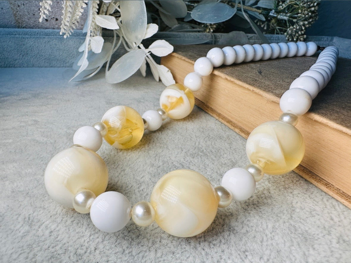 This vintage necklace is a classic. Featuring a graduated design with large, glossy marbled beads in a soft ivory-yellow hue as the focal points. These beads have a semi-translucent quality with swirling patterns that give them a unique appearance. Between the larger marbled beads are smaller, lustrous faux pearl beads and opaque white round beads, adding contrast and a touch of classic sophistication. The beads are symmetrically arranged, with the largest marbled beads positioned centrally, gradually trans