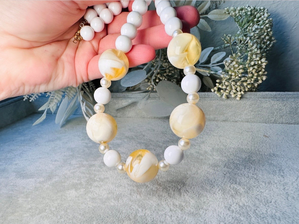 This vintage necklace is a classic. Featuring a graduated design with large, glossy marbled beads in a soft ivory-yellow hue as the focal points. These beads have a semi-translucent quality with swirling patterns that give them a unique appearance. Between the larger marbled beads are smaller, lustrous faux pearl beads and opaque white round beads, adding contrast and a touch of classic sophistication. The beads are symmetrically arranged, with the largest marbled beads positioned centrally, gradually trans