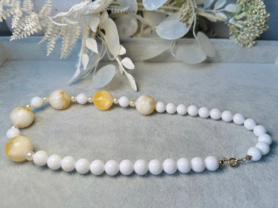 This vintage necklace is a classic. Featuring a graduated design with large, glossy marbled beads in a soft ivory-yellow hue as the focal points. These beads have a semi-translucent quality with swirling patterns that give them a unique appearance. Between the larger marbled beads are smaller, lustrous faux pearl beads and opaque white round beads, adding contrast and a touch of classic sophistication. The beads are symmetrically arranged, with the largest marbled beads positioned centrally, gradually trans
