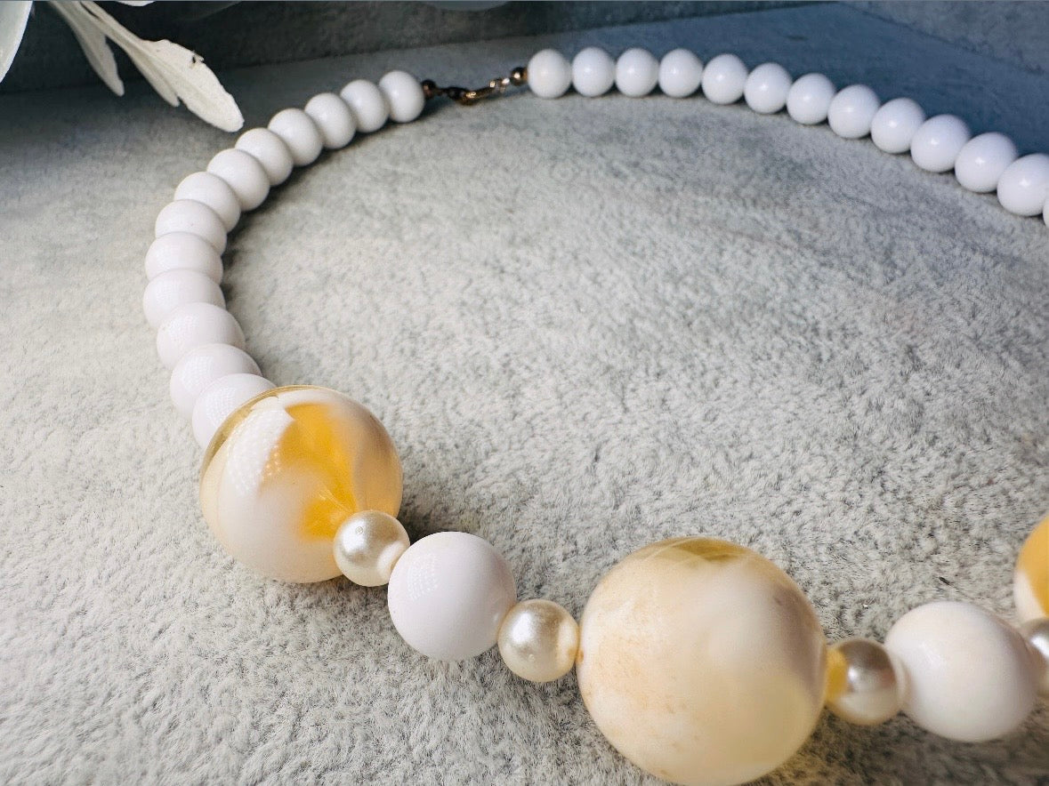 This vintage necklace is a classic. Featuring a graduated design with large, glossy marbled beads in a soft ivory-yellow hue as the focal points. These beads have a semi-translucent quality with swirling patterns that give them a unique appearance. Between the larger marbled beads are smaller, lustrous faux pearl beads and opaque white round beads, adding contrast and a touch of classic sophistication. The beads are symmetrically arranged, with the largest marbled beads positioned centrally, gradually trans