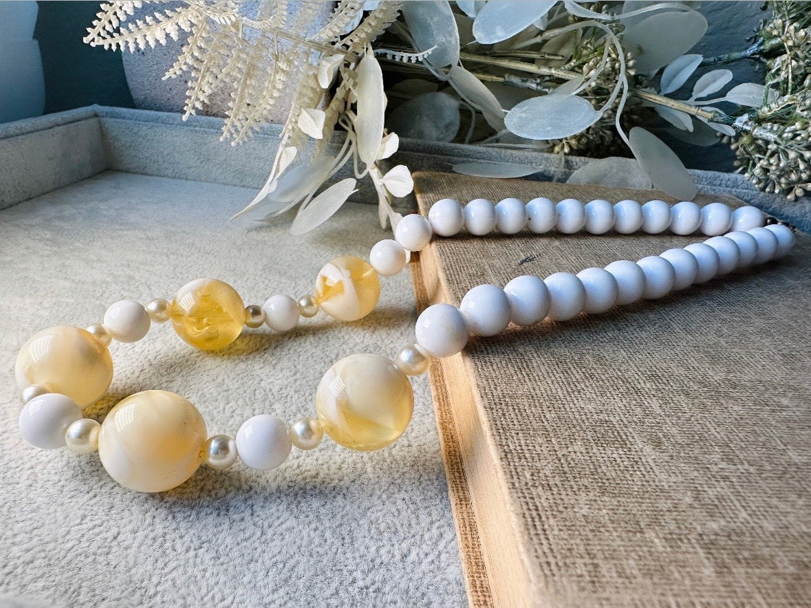 This vintage necklace is a classic. Featuring a graduated design with large, glossy marbled beads in a soft ivory-yellow hue as the focal points. These beads have a semi-translucent quality with swirling patterns that give them a unique appearance. Between the larger marbled beads are smaller, lustrous faux pearl beads and opaque white round beads, adding contrast and a touch of classic sophistication. The beads are symmetrically arranged, with the largest marbled beads positioned centrally, gradually trans