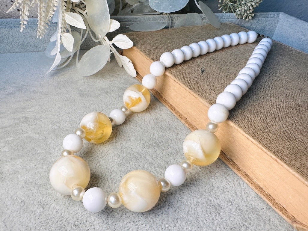 This vintage necklace is a classic. Featuring a graduated design with large, glossy marbled beads in a soft ivory-yellow hue as the focal points. These beads have a semi-translucent quality with swirling patterns that give them a unique appearance. Between the larger marbled beads are smaller, lustrous faux pearl beads and opaque white round beads, adding contrast and a touch of classic sophistication. The beads are symmetrically arranged, with the largest marbled beads positioned centrally, gradually trans