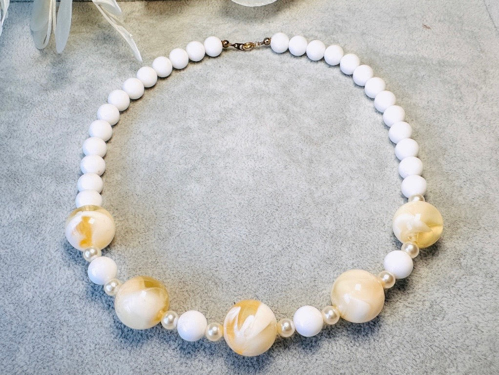 This vintage necklace is a classic. Featuring a graduated design with large, glossy marbled beads in a soft ivory-yellow hue as the focal points. These beads have a semi-translucent quality with swirling patterns that give them a unique appearance. Between the larger marbled beads are smaller, lustrous faux pearl beads and opaque white round beads, adding contrast and a touch of classic sophistication. The beads are symmetrically arranged, with the largest marbled beads positioned centrally, gradually trans