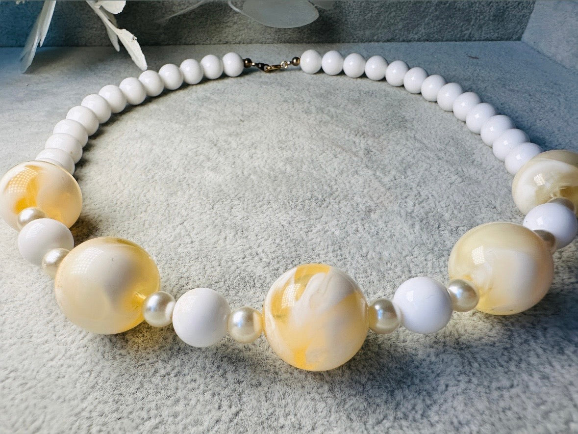 This vintage necklace is a classic. Featuring a graduated design with large, glossy marbled beads in a soft ivory-yellow hue as the focal points. These beads have a semi-translucent quality with swirling patterns that give them a unique appearance. Between the larger marbled beads are smaller, lustrous faux pearl beads and opaque white round beads, adding contrast and a touch of classic sophistication. The beads are symmetrically arranged, with the largest marbled beads positioned centrally, gradually trans