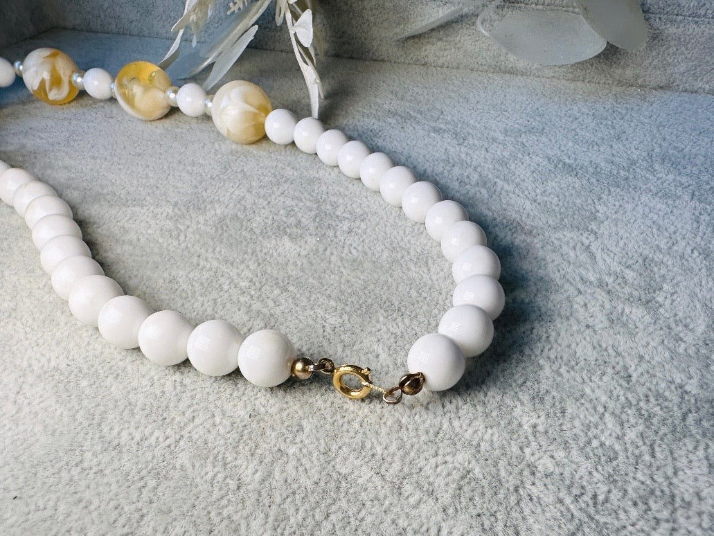 This vintage necklace is a classic. Featuring a graduated design with large, glossy marbled beads in a soft ivory-yellow hue as the focal points. These beads have a semi-translucent quality with swirling patterns that give them a unique appearance. Between the larger marbled beads are smaller, lustrous faux pearl beads and opaque white round beads, adding contrast and a touch of classic sophistication. The beads are symmetrically arranged, with the largest marbled beads positioned centrally, gradually trans