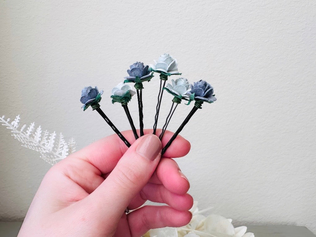 Six handmade rose hair pins with small grey paper flowers. The flowers are in shades of gray; three light silvery grey and three dark charcoal grey. The hair pins are dark bronze. Held in hand for size reference.