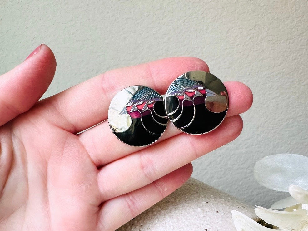 A pair of Laurel Burch vintage earrings titled "Celestial Birds". These post earrings are shiny silver with black and plum pink enamel of three birds lined up in a side profile. Laurel Burch is highly collectible, and these are so unique. Engraved with the the title "Celestial Birds" on the back of earrings along the the signature on the backs. Silver plated posts, excellent condition with very minor wear. They are about 1 inch big and available from Piggle and Pop. held in hand for size reference.