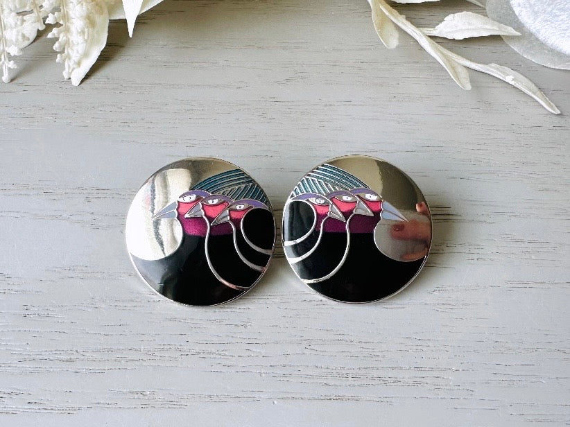 A pair of Laurel Burch vintage earrings titled "Celestial Birds". These post earrings are shiny silver with black and plum pink enamel of three birds lined up in a side profile. Laurel Burch is highly collectible, and these are so unique. Engraved with the the title "Celestial Birds" on the back of earrings along the the signature on the backs. Silver plated posts, excellent condition with very minor wear. They are about 1 inch big and available from Piggle and Pop.