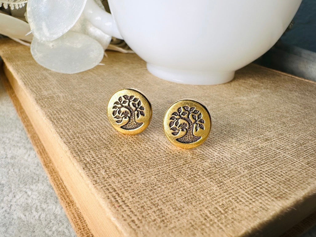 Gold tree-of-life earrings on a wooden surface with a white teapot in the background.