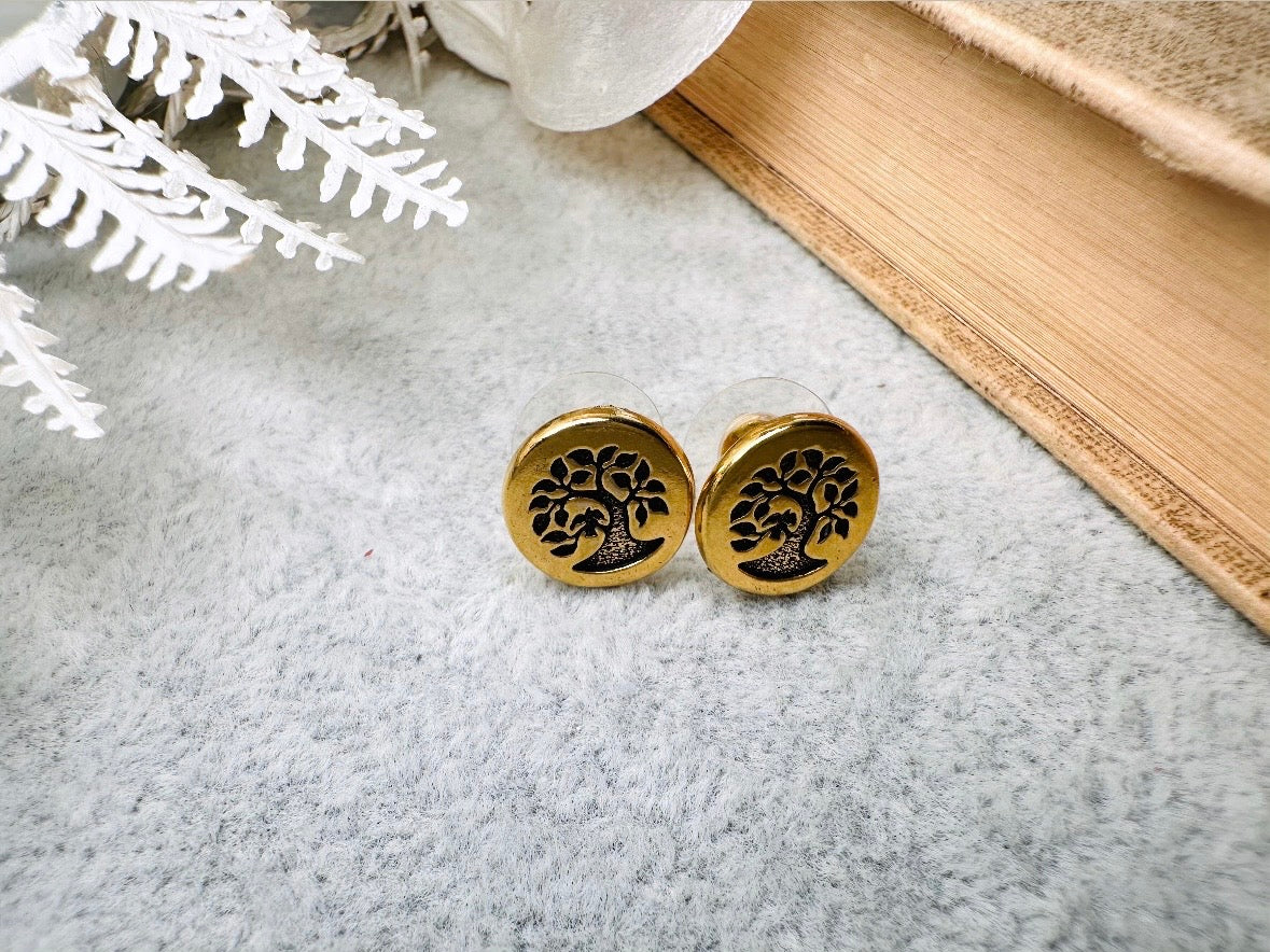 Gold tree-of-life earrings on a textured surface with a book and decorative branches in the background.