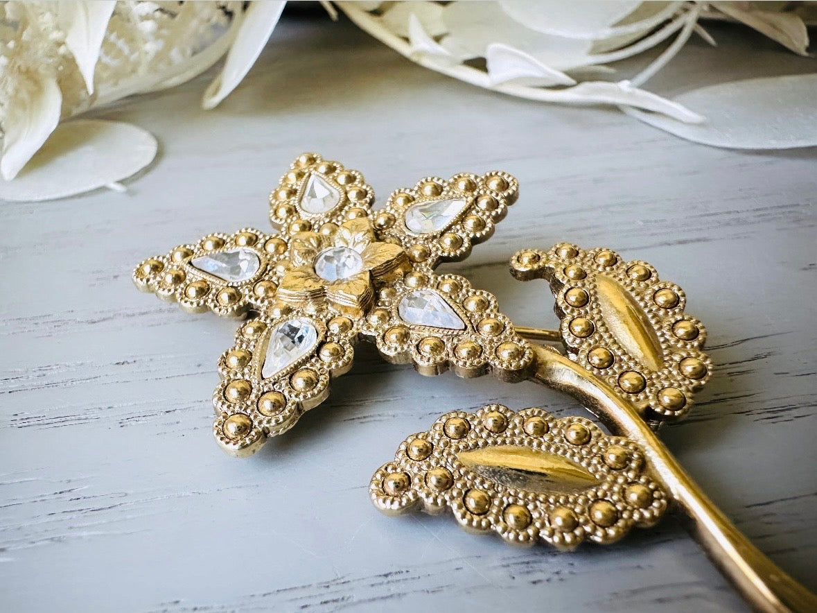 A beautiful vintage flower brooch by the brand 1928. The brooch is a single flower shape that is gold, with textured dotting all around each petal and leaf. There are diamond crystal rhinestones in a teardrop shape in the center of each flower petal and a round crystal in the center. The stem is a smooth shiny gold. The brooch is on the signature background of the shop Piggle and Pop, a light gray wood table with white floral background. This is a close up of the details.