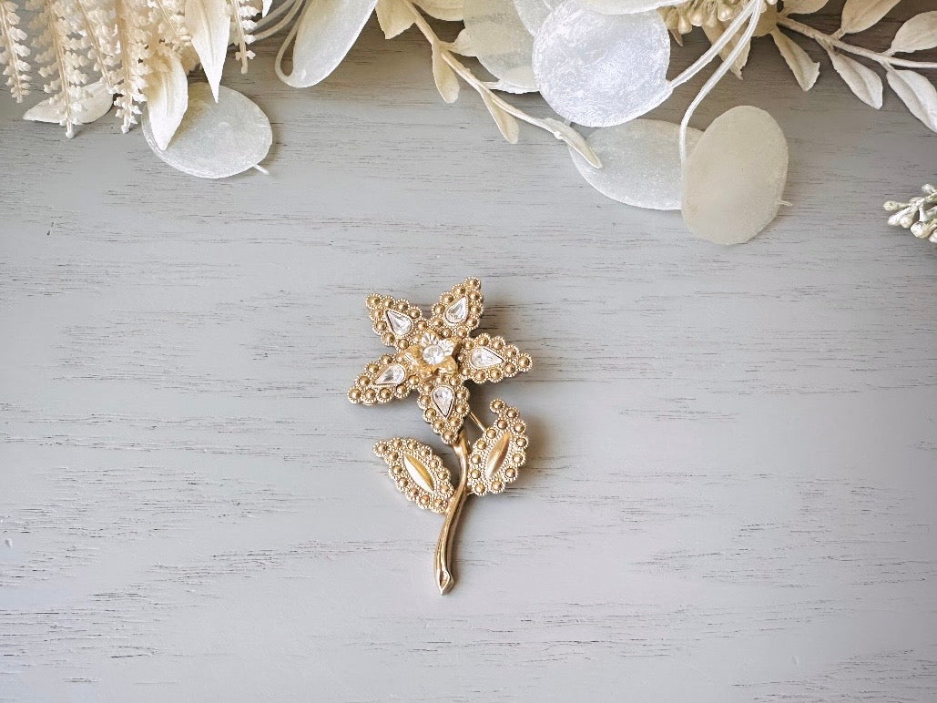 A beautiful vintage flower brooch by the brand 1928. The brooch is a single flower shape that is gold, with textured dotting all around each petal and leaf. There are diamond crystal rhinestones in a teardrop shape in the center of each flower petal and a round crystal in the center. The stem is a smooth shiny gold. The brooch is on the signature background of the shop Piggle and Pop, a light gray wood table with white floral background.