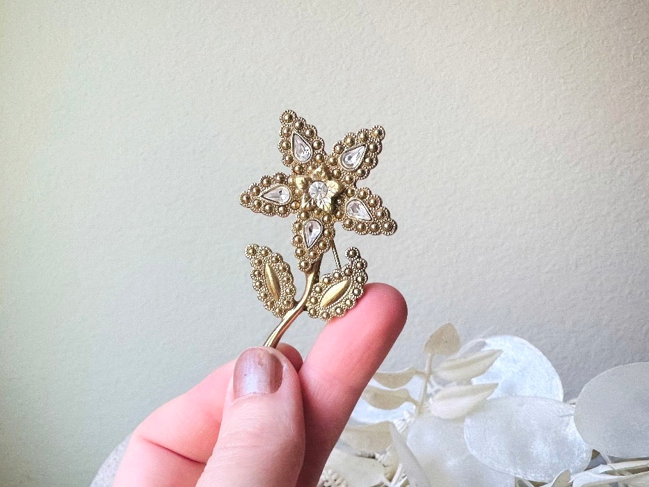 A beautiful vintage flower brooch by the brand 1928. The brooch is a single flower shape that is gold, with textured dotting all around each petal and leaf. There are diamond crystal rhinestones in a teardrop shape in the center of each flower petal and a round crystal in the center. The stem is a smooth shiny gold. The brooch is on the signature background of the shop Piggle and Pop, a light gray wood table with white floral background. Held in hand for size reference.