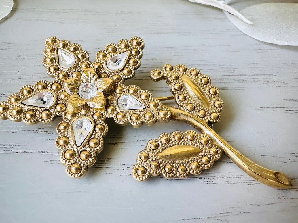 A beautiful vintage flower brooch by the brand 1928. The brooch is a single flower shape that is gold, with textured dotting all around each petal and leaf. There are diamond crystal rhinestones in a teardrop shape in the center of each flower petal and a round crystal in the center. The stem is a smooth shiny gold. The brooch is on the signature background of the shop Piggle and Pop, a light gray wood table with white floral background.
