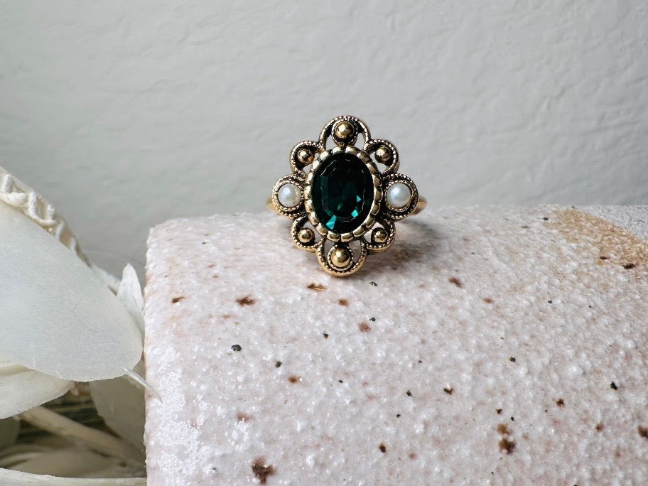 This vintage emerald green and pearl gold cocktail ring is such a pretty piece. Vintage Avon from the 1976 Versailles collection. Green, pearls and gold are a beautiful color combination. The center faceted oval green stone is surrounded in a raised filigree design that is full of vintage character, with two tiny pearl cabochon accents. Signed in excellent vintage condition. 