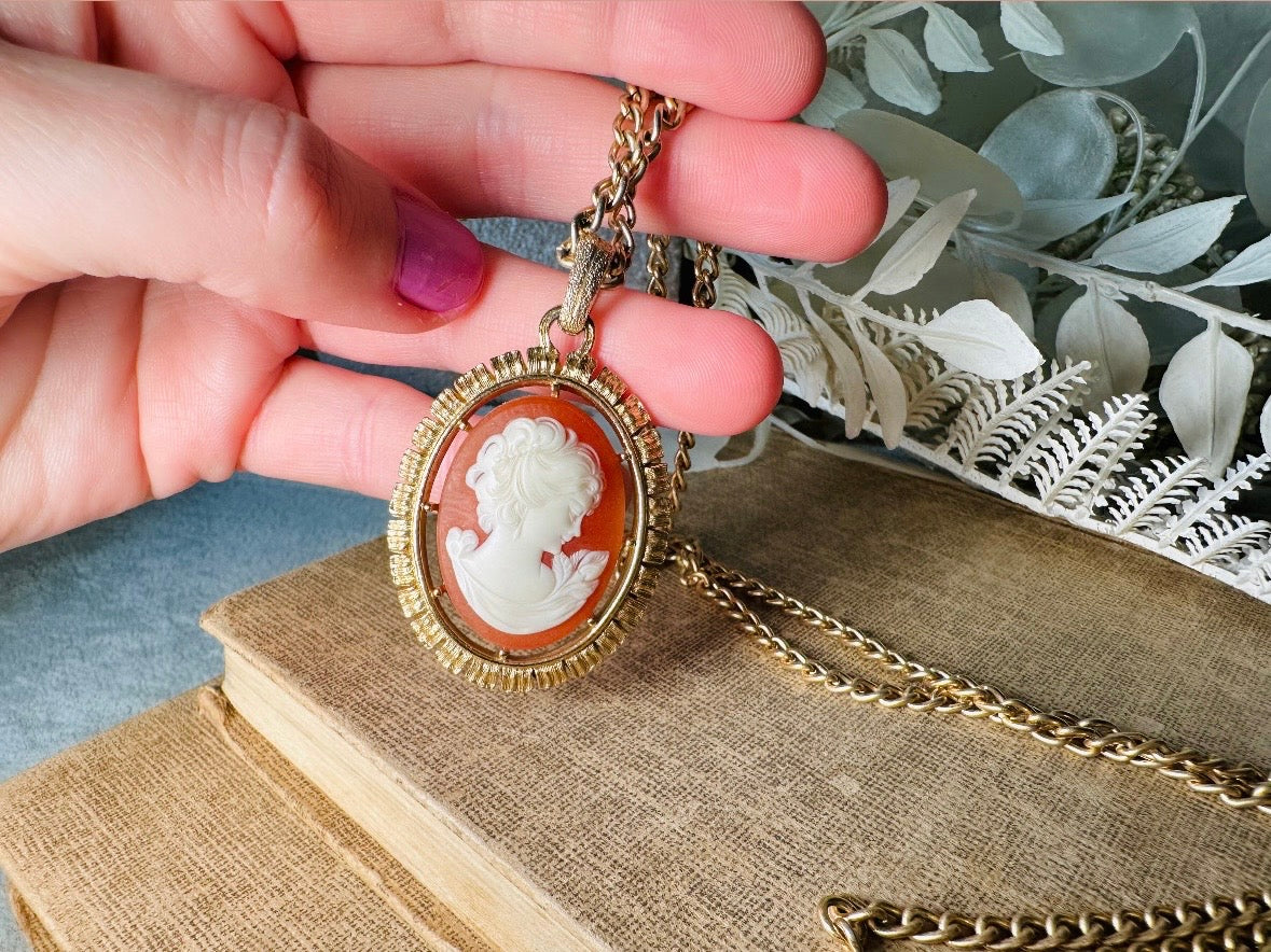 This 1970s vintage Victorian Revival cameo necklace is marvelous. Bold, yet elegant this classic necklace features a double sided reversible cameo pendant. On one side is the portrait of a lady in cream and classic muted orange and on the other side an ornate Fleur de Lis raised in gold. This vintage necklace features a medium width antique gold curb chain, from 1974. In great vintage condition please view all pics. Would look incredible in a necklace stack for a modern twist.