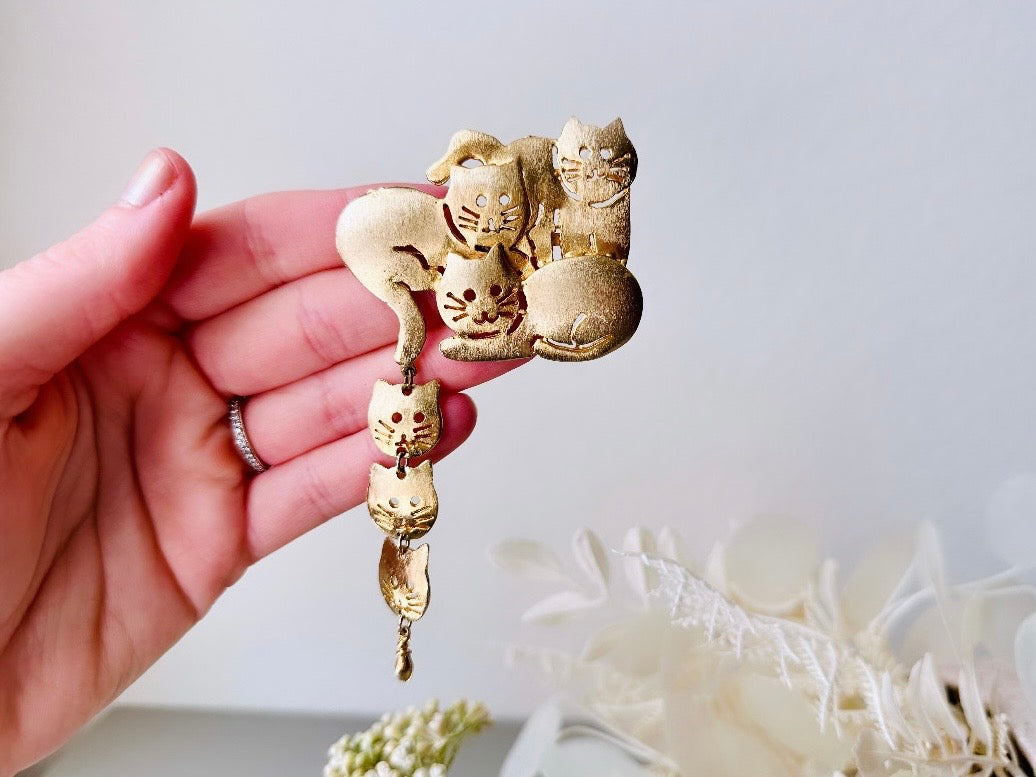 This is a big gold vintage 1980s cat brooch featuring three cats huddled together and three cat heads dangling from their tail. The gold is a classic semi- matte shade with brushed detailing. It is a signed Ultra Craft piece in excellent vintage condition.