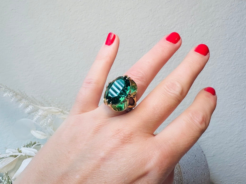 Hand wearing a large green gemstone ring with red nail polish on a light gray background