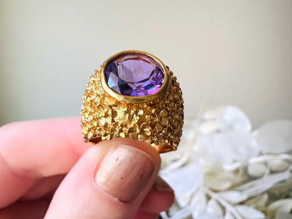 A vintage 1970s cocktail ring, that is gold and purple, The gold band has a raised textured gold nugget surface with a beautiful faceted round cut vibrant amethyst purple in the center.