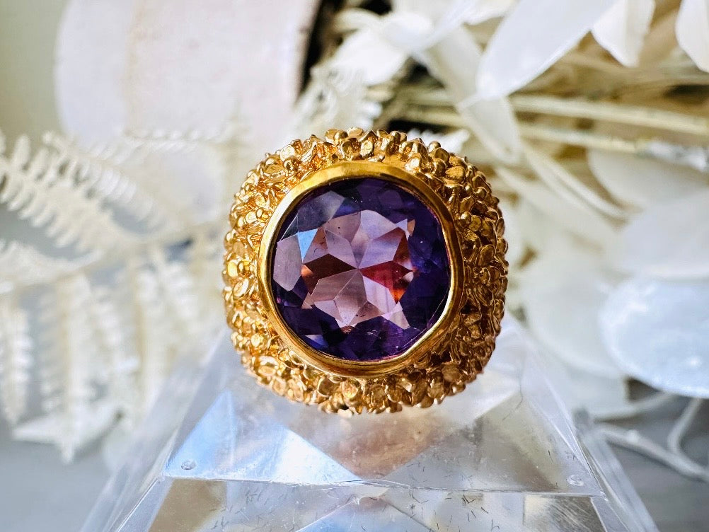 A vintage 1970s cocktail ring, that is gold and purple, The gold band has a raised textured gold nugget surface with a beautiful faceted round cut vibrant amethyst purple in the center. Close up Shot