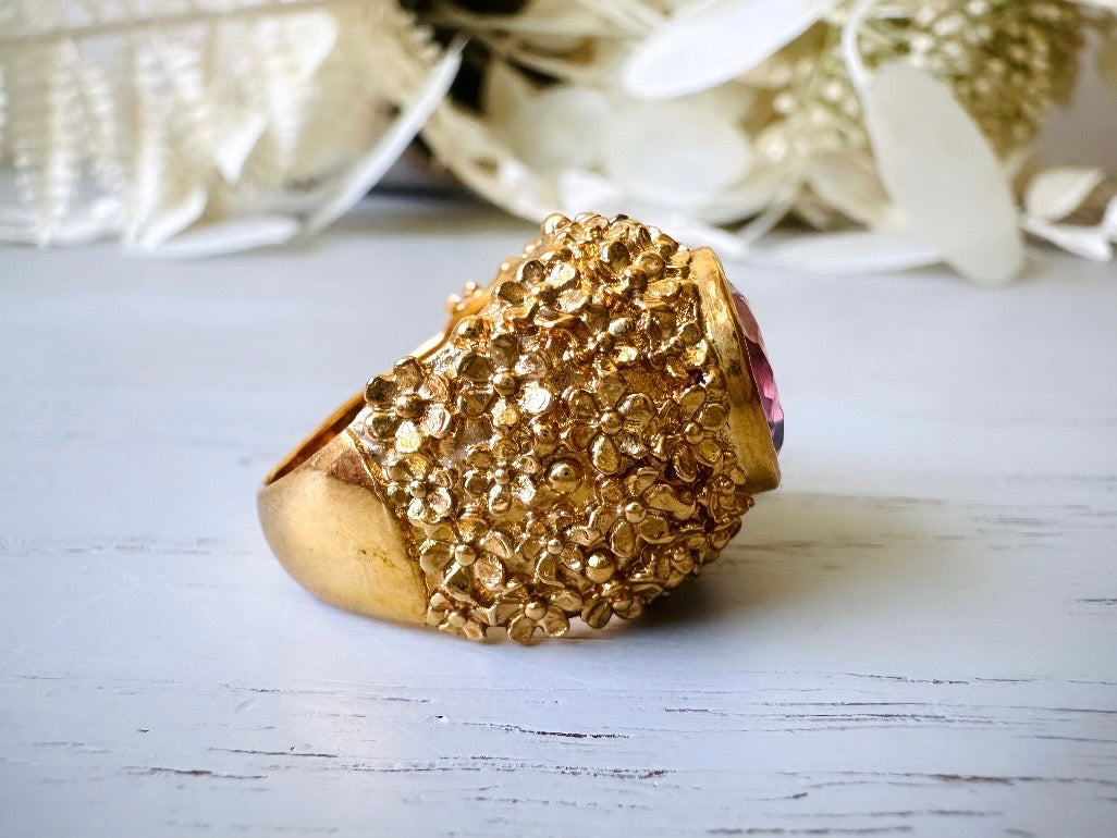 A vintage 1970s cocktail ring, that is gold and purple, The gold band has a raised textured gold nugget floral surface with a beautiful faceted round cut vibrant amethyst purple in the center. Side view