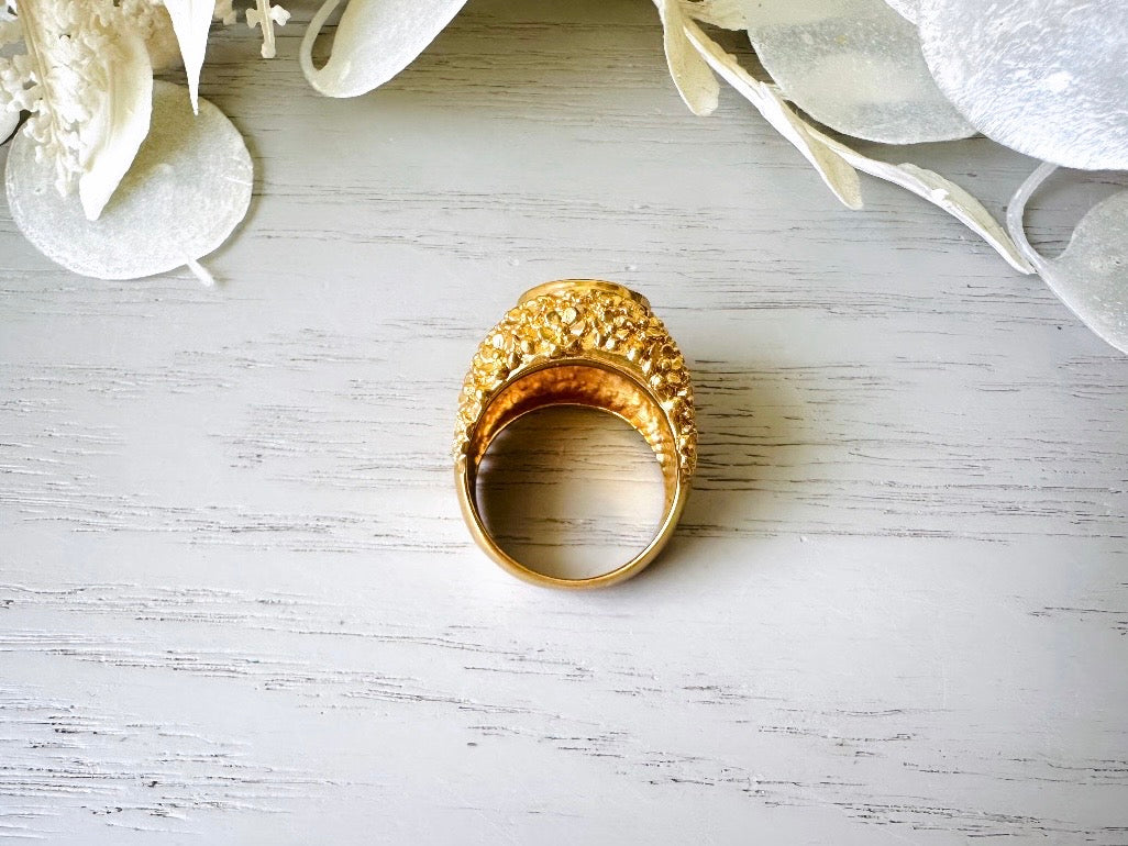 A vintage 1970s cocktail ring, that is gold and purple, The gold band has a raised textured gold nugget surface with a beautiful faceted round cut vibrant amethyst purple in the center.