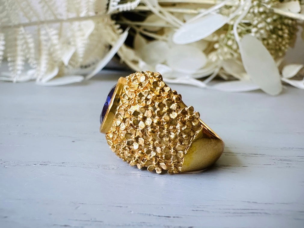 A vintage 1970s cocktail ring, that is gold and purple, The gold band has a raised textured gold nugget surface with a beautiful faceted round cut vibrant amethyst purple in the center.