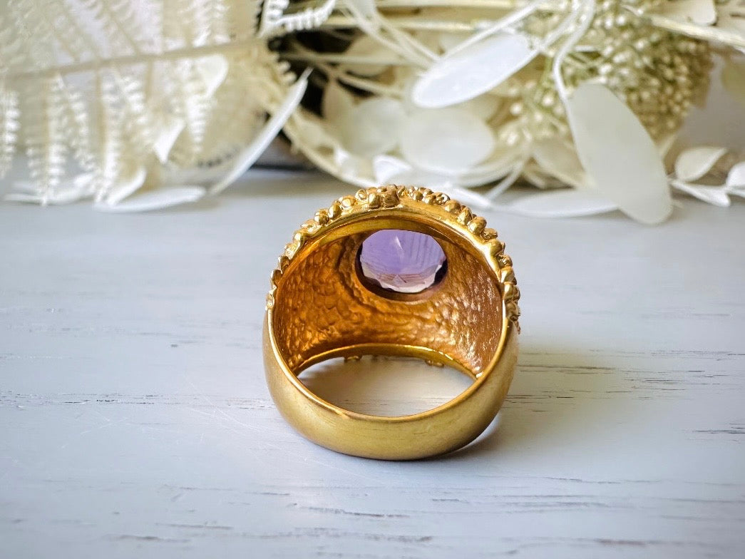A vintage 1970s cocktail ring, that is gold and purple, The gold band has a raised textured gold nugget surface with a beautiful faceted round cut vibrant amethyst purple in the center.