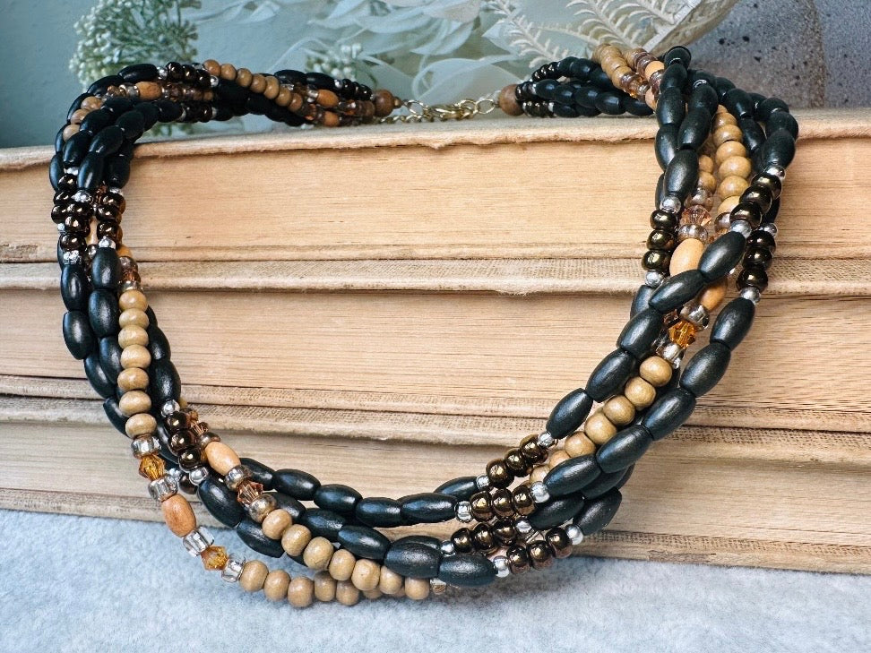 This vintage Napier necklace is a beautiful beaded twisted multistrand choker. Featuring six beaded strands in varying shapes and sizes composed of beads in wood, glass, acrylic, this bold necklace is a fabulous addition to your jewelry box. The beaded strands connect with a gold extension chain finished with a beautiful stamped Napier tag. Earthy black, brown and cream wood paired with gold, bronze and clear crystals- this necklace pairs beautifully with a variety of earrings and bracelets. Minimal wear in