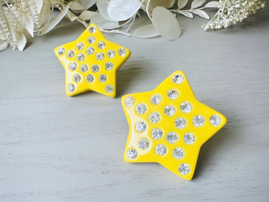 A pair of yellow star earrings from Piggle and Pop. A highly collectible pair of 1980s statement earrings. These yellow star earrings are bright banana yellow opaque lucite with diamond rhinestone accents. Huge vintage star clip ons with major rockstar vibes. close up