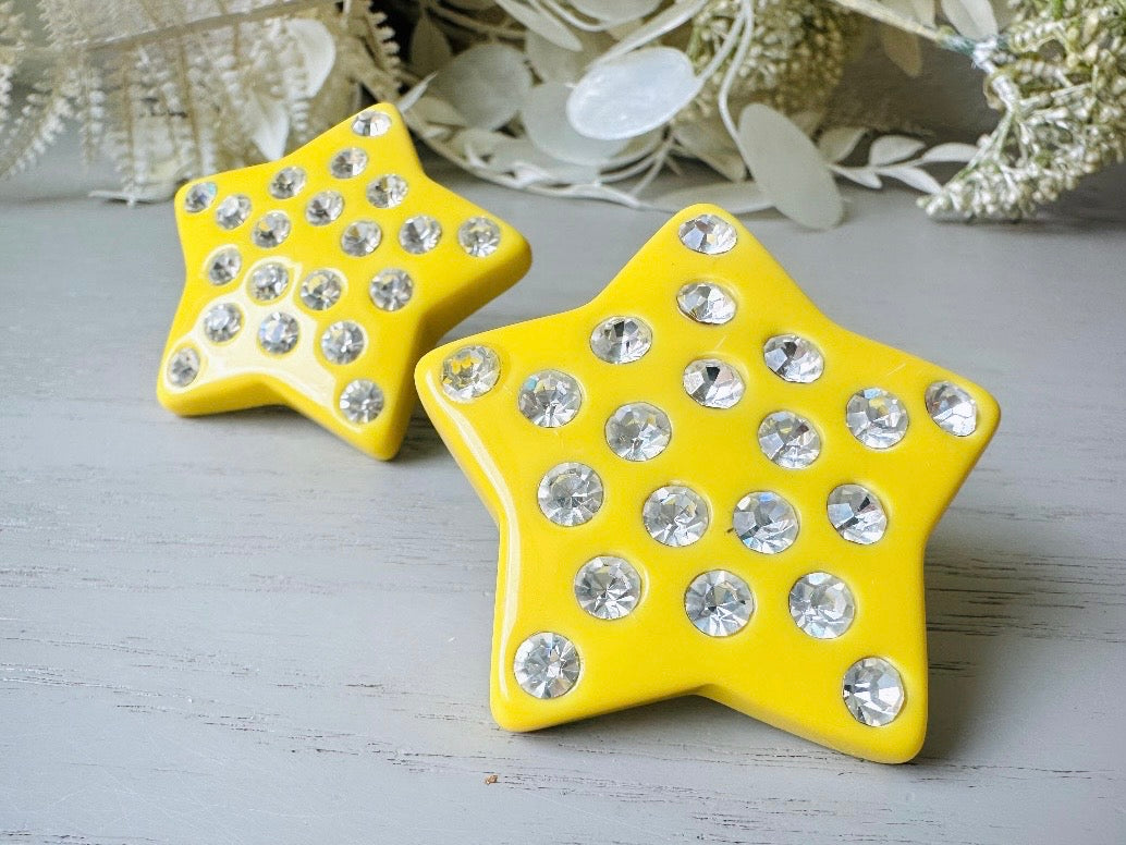 A pair of yellow star earrings from Piggle and Pop. A highly collectible pair of 1980s statement earrings. These yellow star earrings are bright banana yellow opaque lucite with diamond rhinestone accents. Huge vintage star clip ons with major rockstar vibes. super close up