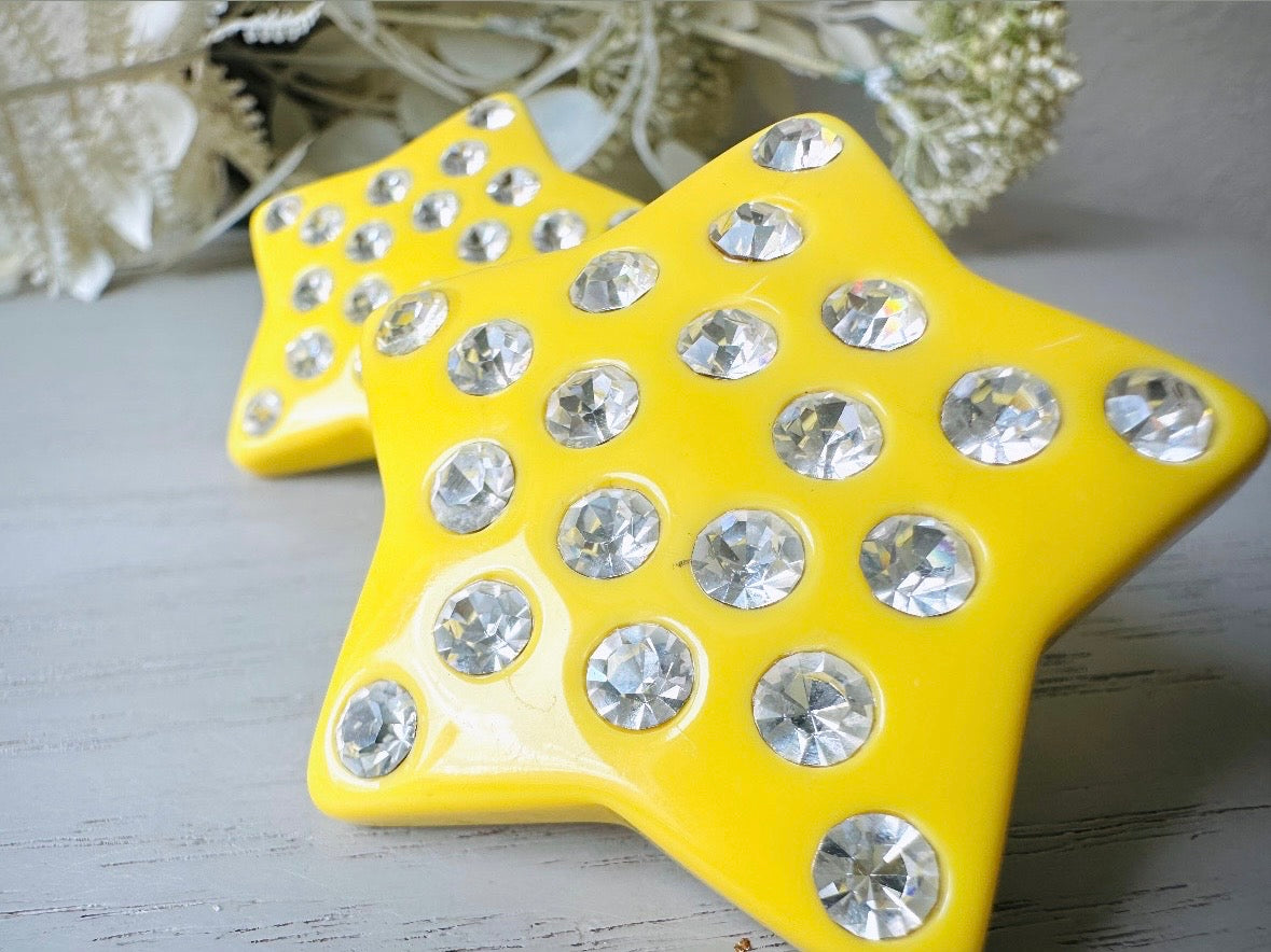 A pair of yellow star earrings from Piggle and Pop. A highly collectible pair of 1980s statement earrings. These yellow star earrings are bright banana yellow opaque lucite with diamond rhinestone accents. Huge vintage star clip ons with major rockstar vibes. extreme close up