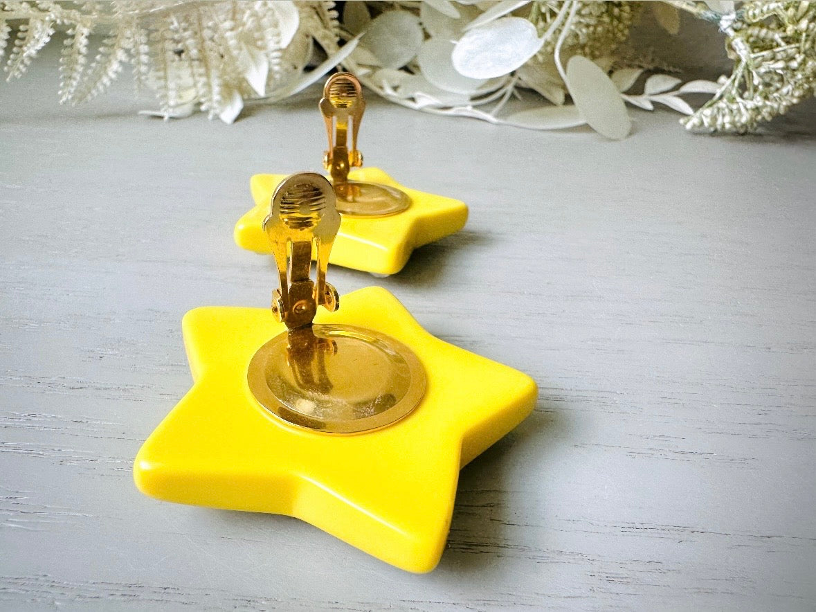 A pair of yellow star earrings from Piggle and Pop. A highly collectible pair of 1980s statement earrings. These yellow star earrings are bright banana yellow opaque lucite with diamond rhinestone accents. Huge vintage star clip ons with major rockstar vibes. backs of earring paddles