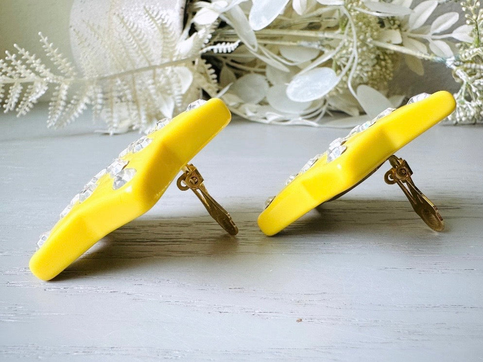 A pair of yellow star earrings from Piggle and Pop. A highly collectible pair of 1980s statement earrings. These yellow star earrings are bright banana yellow opaque lucite with diamond rhinestone accents. Huge vintage star clip ons with major rockstar vibes. side view.