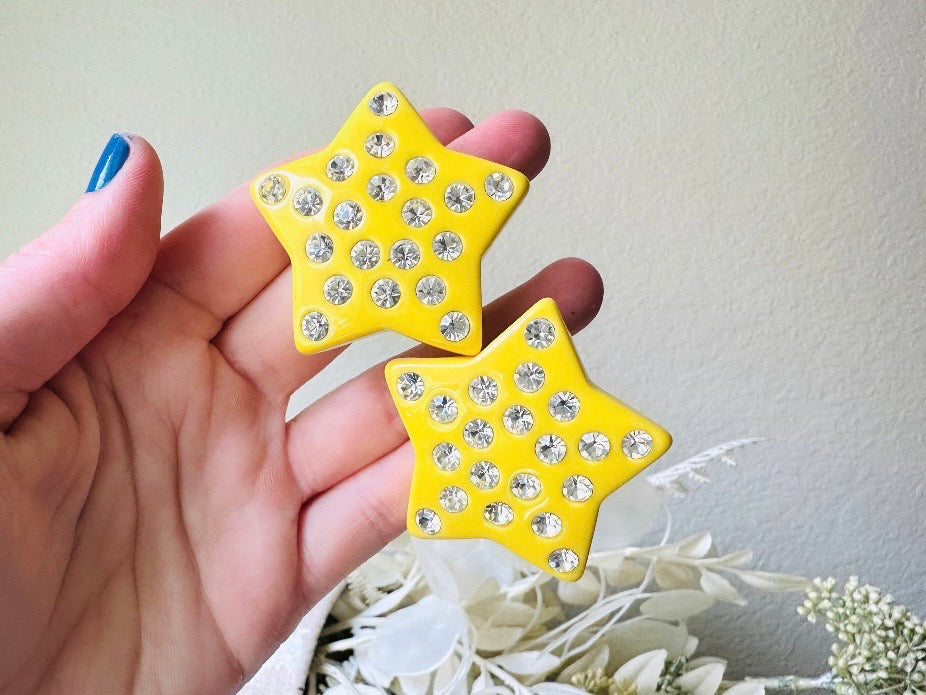 A pair of yellow star earrings from Piggle and Pop. A highly collectible pair of 1980s statement earrings. These yellow star earrings are bright banana yellow opaque lucite with diamond rhinestone accents. Huge vintage star clip ons with major rockstar vibes. held in hand for size reference.