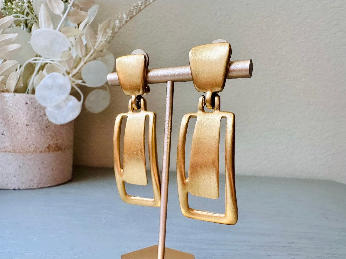 These Anne Klein vintage matte gold designer earrings are spectacular. Full of 1980s bold glamour, these clip ons have a satin gold finish and a geometric design. These vintage dangle earrings are bold and elegant, with curved square posts and rectangular drops with a cut out hollow border. These are big and incredibly well made in a satin matte gold finish. Clip on style, easy to wear and in great vintage condition. Highly sought after designer vintage. 