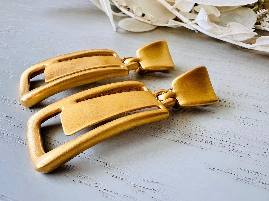 These Anne Klein vintage matte gold designer earrings are spectacular. Full of 1980s bold glamour, these clip ons have a satin gold finish and a geometric design. These vintage dangle earrings are bold and elegant, with curved square posts and rectangular drops with a cut out hollow border. These are big and incredibly well made in a satin matte gold finish. Clip on style, easy to wear and in great vintage condition. Highly sought after designer vintage. 