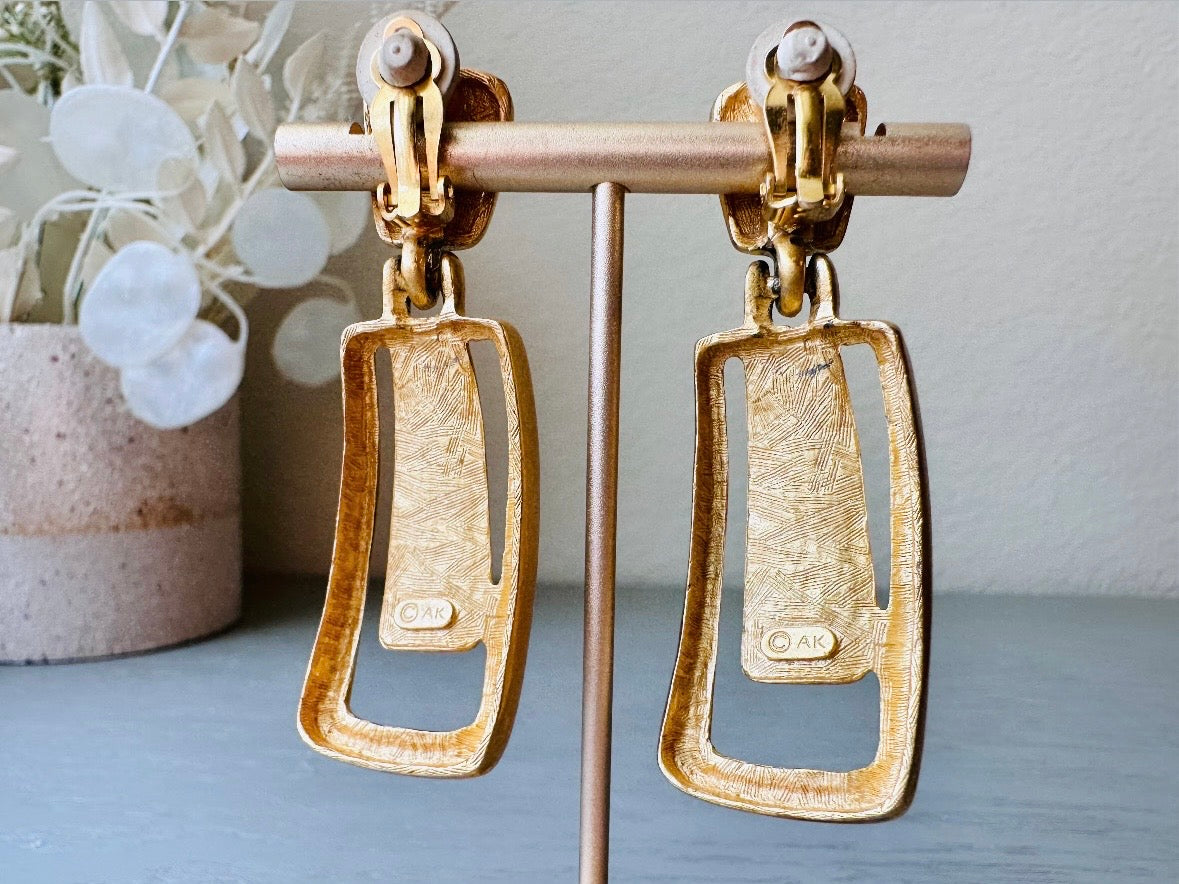 These Anne Klein vintage matte gold designer earrings are spectacular. Full of 1980s bold glamour, these clip ons have a satin gold finish and a geometric design. These vintage dangle earrings are bold and elegant, with curved square posts and rectangular drops with a cut out hollow border. These are big and incredibly well made in a satin matte gold finish. Clip on style, easy to wear and in great vintage condition. Highly sought after designer vintage. 