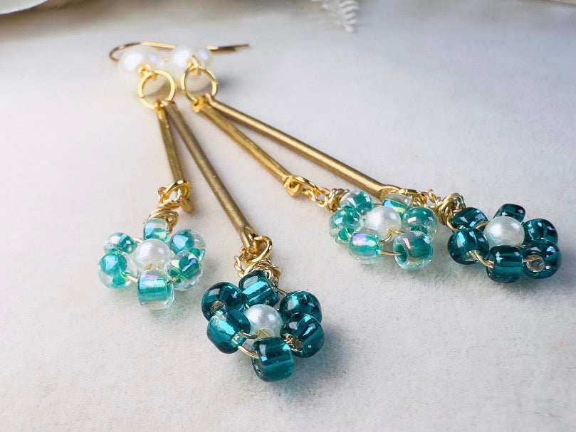 A pair of handmade dangle earrings with gold-tone metal rods, small white pearl beads, and clusters of teal and aqua blue faceted beads, displayed on a soft white textured surface.