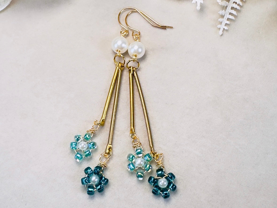 A pair of handmade dangle earrings with gold-tone metal, white pearl beads, and teal blue seed beads arranged in a floral pattern, displayed on a light beige textured surface.