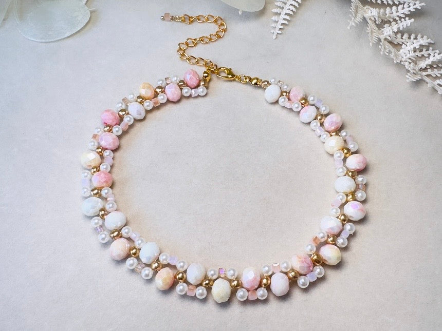 A delicate beaded necklace with pastel pink, white, and peach beads, accented by small pearl-like beads and gold-tone metal links and clasp, displayed on a light beige textured surface.
