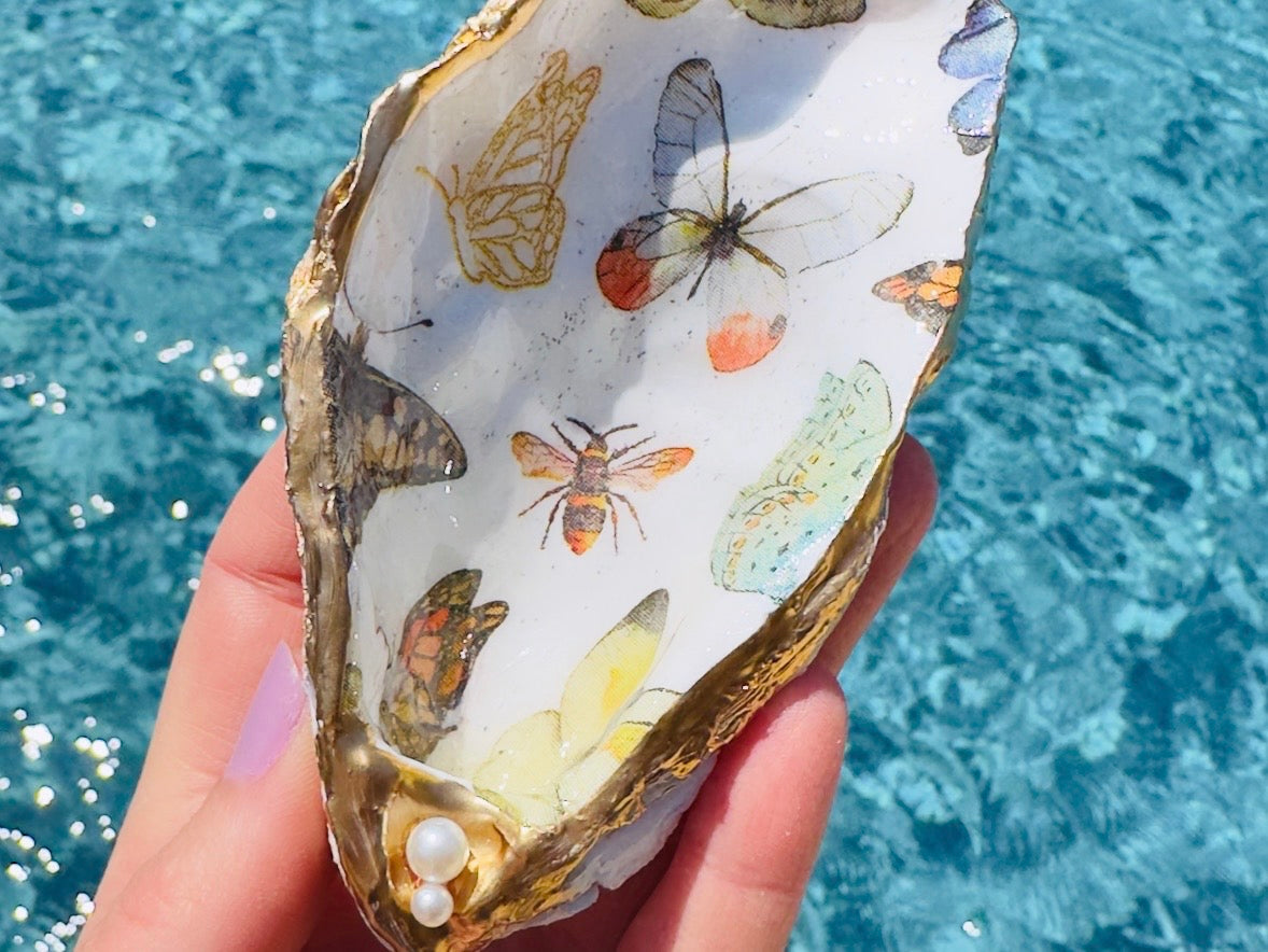 A hand holds an open oyster shell decorated with painted butterflies and bees, set against a bright blue swimming pool background.