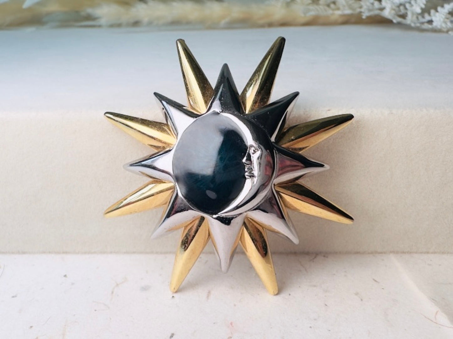 A two-tone metal sun and moon brooch with a polished silver crescent moon face and alternating gold and silver star points, displayed on a light beige textured surface.