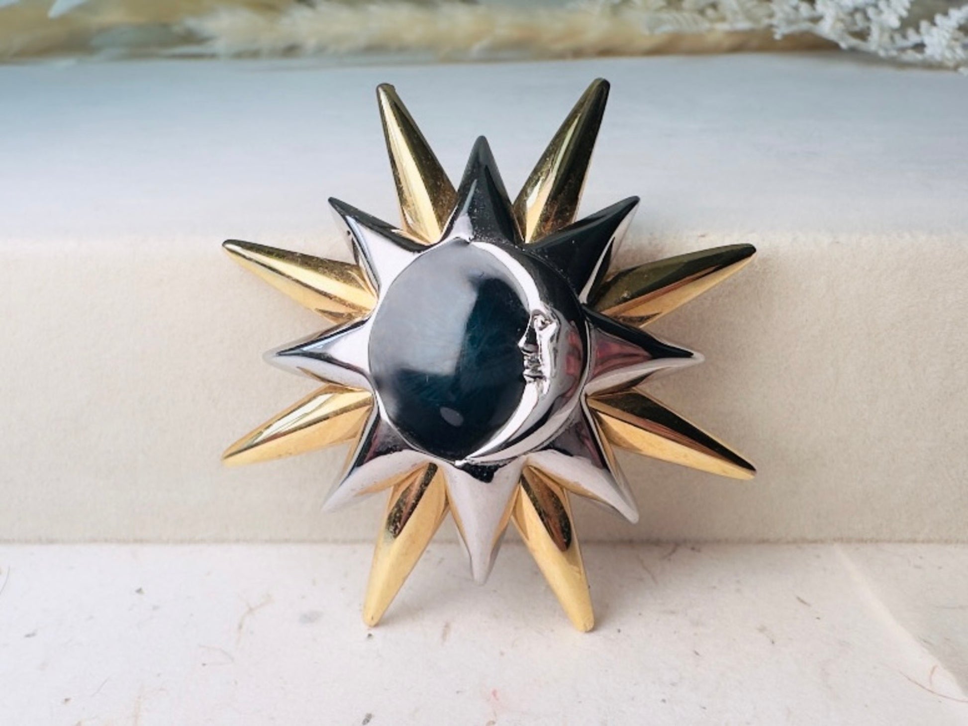 A two-tone metal sun and moon brooch with a polished silver crescent moon face and alternating gold and silver star points, displayed on a light beige textured surface.