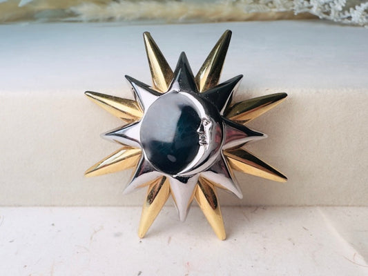 A two-tone metal sun and moon brooch with a polished silver crescent moon face and alternating gold and silver star points, displayed on a light beige textured surface.