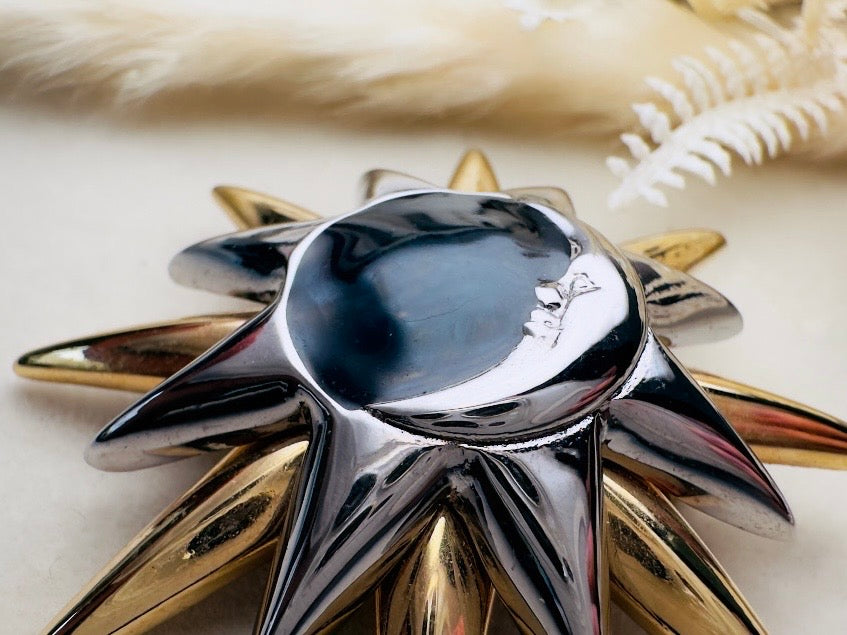 A close-up of a metallic starburst-shaped decorative object with a polished silver center and alternating gold and silver rays, photographed on a light neutral surface with soft dried floral props in the background.