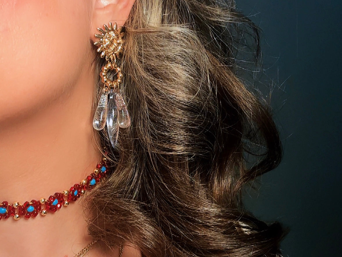 A close-up of a model's ear wearing a large ornate gold-toned statement earring with clear teardrop crystals, alongside a red and turquoise beaded choker necklace.