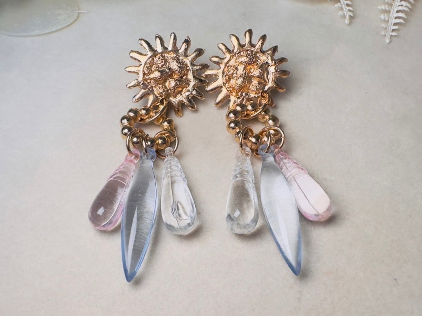 A pair of dangling statement earrings with textured gold sunburst tops and three teardrop-shaped clear and pale pink glass drops, displayed on a light beige surface.